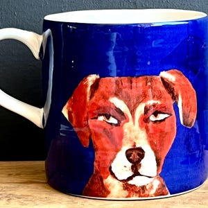 May include: A ceramic mug with a white handle and interior. The exterior is a deep blue with a painted illustration of a dog's face in shades of red, brown, and white. The dog has a slightly grumpy expression.