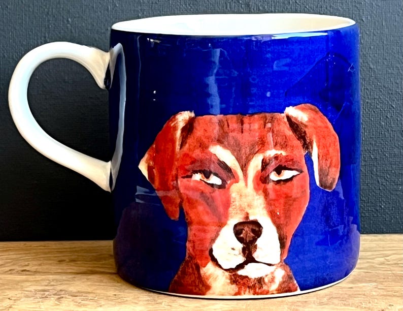 May include: A ceramic mug with a white handle and interior. The exterior is a deep blue with a painted illustration of a dog's face in shades of red, brown, and white. The dog has a slightly grumpy expression.