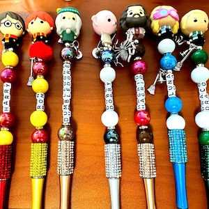 May include: Seven pens topped with character figures including Harry, Ron, Dumbledore, Dobby, Hagrid, Luna, and Draco. Each pen features colorful beads, lettered cubes spelling the character names, and a sparkling rhinestone barrel.