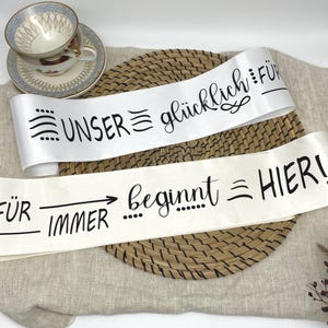 Könnte beinhalten: Zwei weiße Satinbänder mit schwarzer Schrift. Auf dem oberen Band steht „UNSER glücklich FÜR“ und auf dem unteren Band „FÜR immer beginnt HIER!“. Eine dekorative Teetasse und Untertasse sind im Hintergrund.