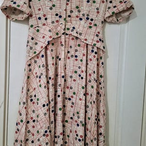 May include: A light beige dress featuring a pattern of red, green, and dark blue polka dots overlaid with thin vertical and horizontal red lines. The dress has short sleeves with cuffs, a pointed collar, and a front zip closure extending to the waist.