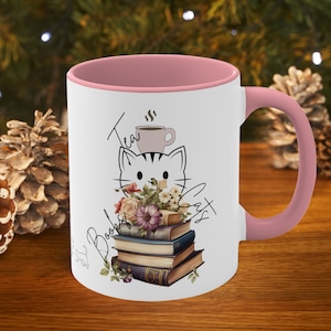 May include: White ceramic mug with a pink rim and handle. The mug features a black and white illustration of a cat sitting on a stack of books with flowers. The text "Tea, Books & Cats" is written on the mug.