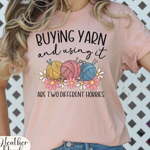 Puede incluir: Camiseta rosa claro con el texto "BUYING YARN and using it ARE TWO DIFFERENT HOBBIES". El dise&ntilde;o presenta tres ovillos de lana en amarillo, rosa y azul, con margaritas.