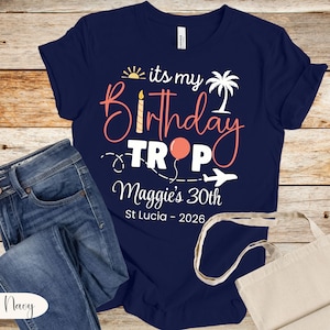May include: Navy blue t-shirt with the text "its my Birthday Trop Maggie's 30th St Lucia - 2026" in a tropical design. The design includes a sun, palm tree, balloon, and airplane. The shirt is paired with blue jeans.