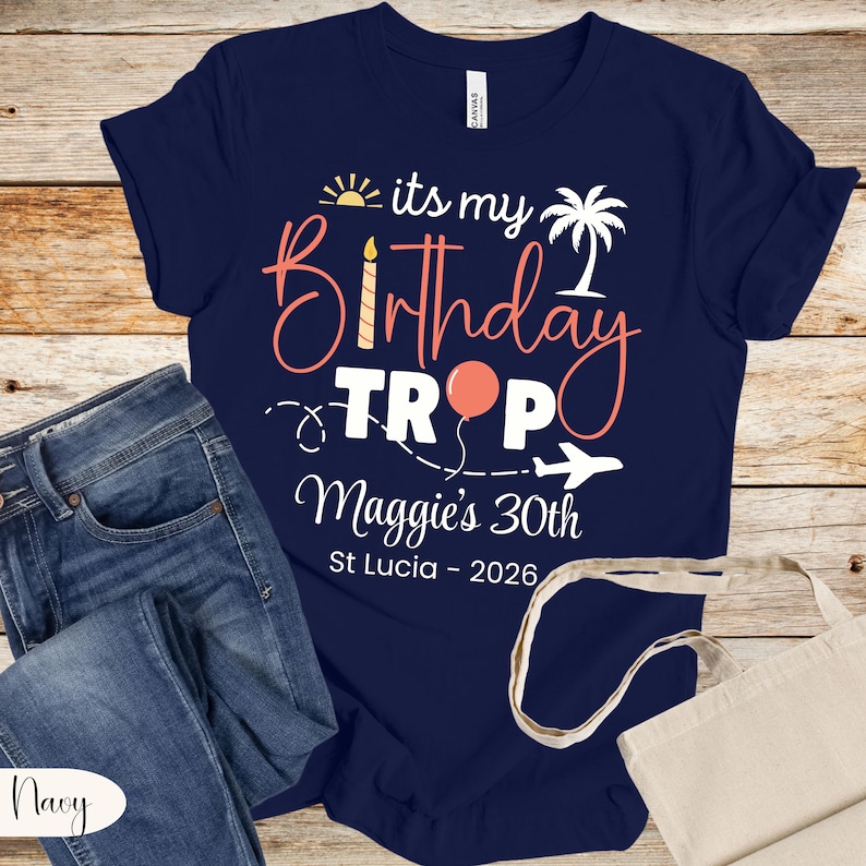 May include: Navy blue t-shirt with the text "its my Birthday Trop Maggie's 30th St Lucia - 2026" in a tropical design. The design includes a sun, palm tree, balloon, and airplane. The shirt is paired with blue jeans.