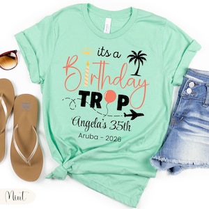 May include: Mint green t-shirt with the text "its a Birthday Trip Angela's 35th Aruba - 2026". The design includes a sun, palm tree, balloon, and airplane. The shirt is paired with denim shorts and sandals.