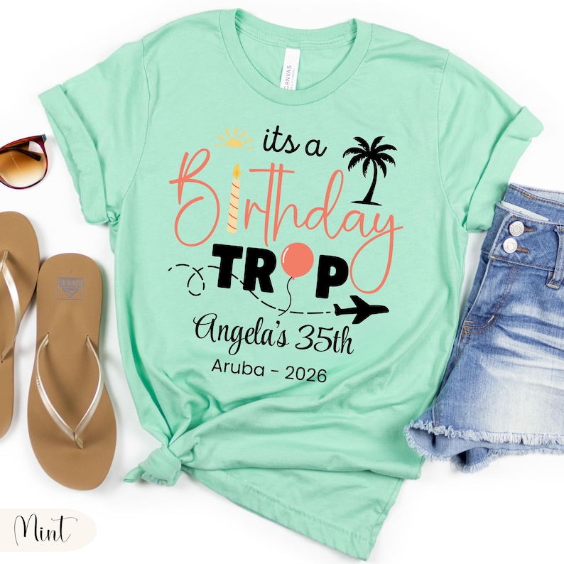 May include: Mint green t-shirt with the text "its a Birthday Trip Angela's 35th Aruba - 2026". The design includes a sun, palm tree, balloon, and airplane. The shirt is paired with denim shorts and sandals.