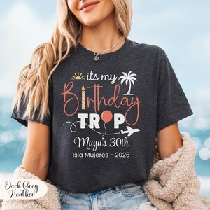May include: Dark grey heather t-shirt with the text "its my Birthday Trop" in coral and white. The design includes a sun, palm tree, balloon, and airplane. The text "Maya's 30th Isla Mujeres - 2026" is also printed on the shirt.
