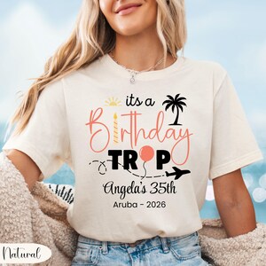May include: Cream-colored t-shirt with the text "its a Birthday Trip" in pink and black. The design includes a sun, palm tree, balloon, and airplane. Below, it reads "Angela's 35th Aruba - 2026".
