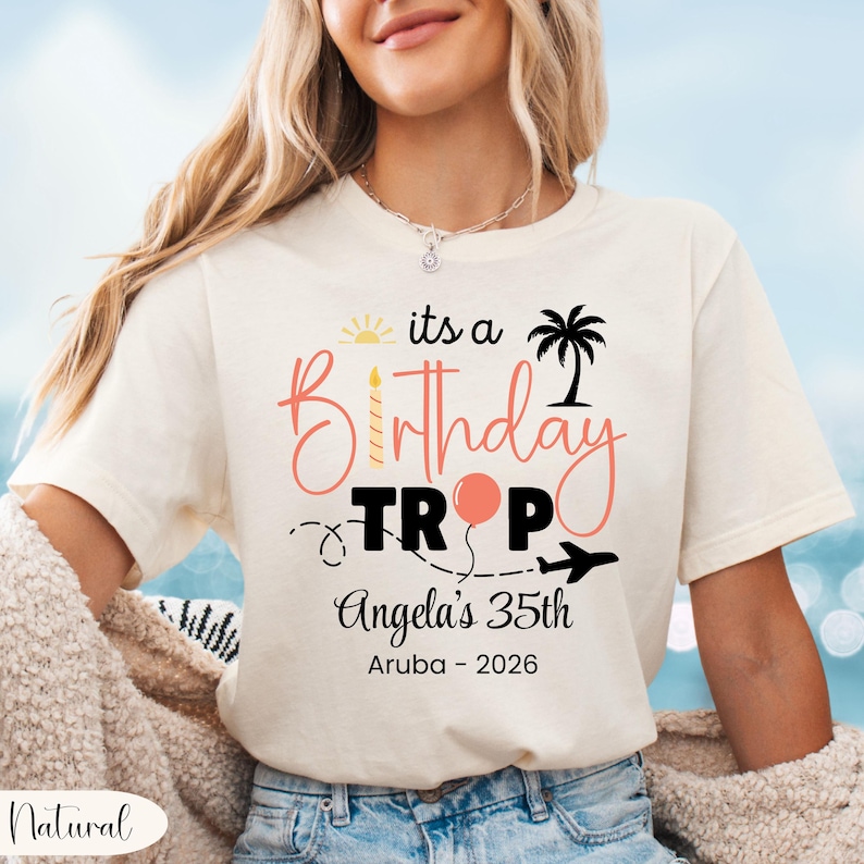 May include: Cream-colored t-shirt with the text "its a Birthday Trip" in pink and black. The design includes a sun, palm tree, balloon, and airplane. Below, it reads "Angela's 35th Aruba - 2026".