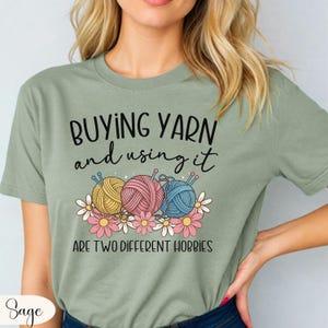 Puede incluir: Camiseta verde salvia con el texto "COMPRAR HILO y usarlo SON DOS HOBBIES DIFERENTES". La camiseta presenta ilustraciones de ovillos de lana en amarillo, rosa y azul, con agujas de tejer y flores. Una camiseta divertida para artesanos.