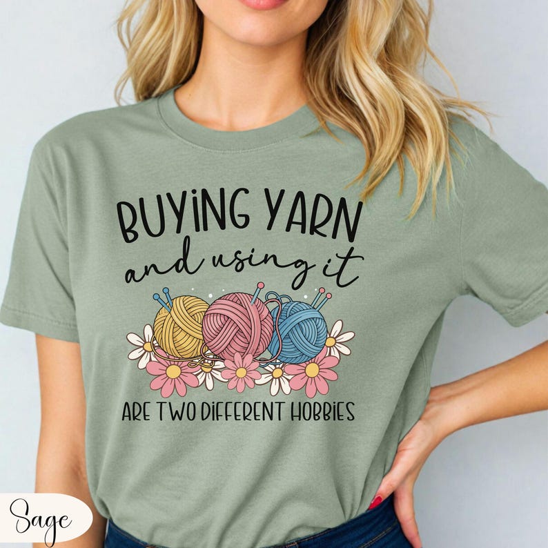 Puede incluir: Camiseta verde salvia con el texto "COMPRAR HILO y usarlo SON DOS HOBBIES DIFERENTES". La camiseta presenta ilustraciones de ovillos de lana en amarillo, rosa y azul, con agujas de tejer y flores. Una camiseta divertida para artesanos.