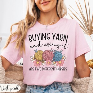 Puede incluir: Camiseta rosa claro con el texto "BUYING YARN and using it ARE TWO DIFFERENT HOBBIES". La camiseta presenta un gr&aacute;fico de tres ovillos de lana en amarillo, rosa y azul, con agujas de tejer y flores.