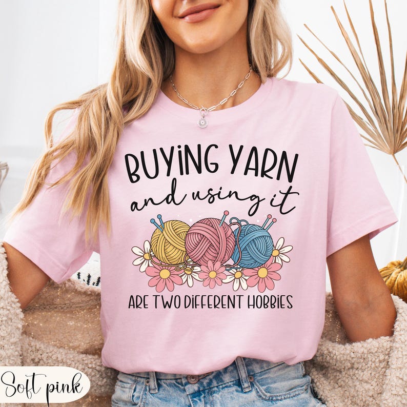 Puede incluir: Camiseta rosa claro con el texto "BUYING YARN and using it ARE TWO DIFFERENT HOBBIES". La camiseta presenta un gr&aacute;fico de tres ovillos de lana en amarillo, rosa y azul, con agujas de tejer y flores.