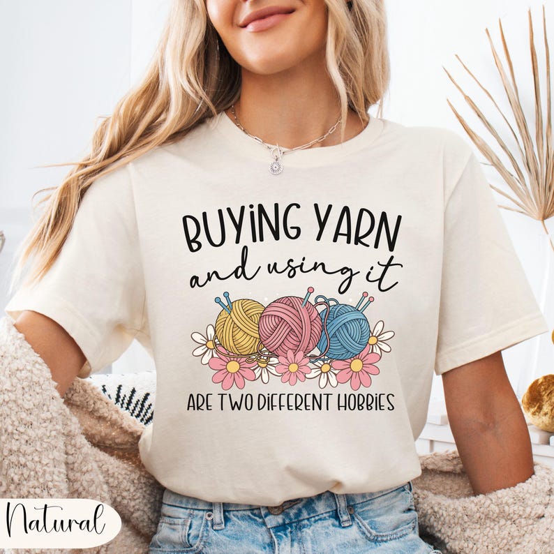 Puede incluir: Camiseta color crema con el texto "BUYING YARN and using it ARE TWO DIFFERENT HOBBIES." El gr&aacute;fico presenta tres ovillos de lana en amarillo, rosa y azul, con agujas de tejer y flores.