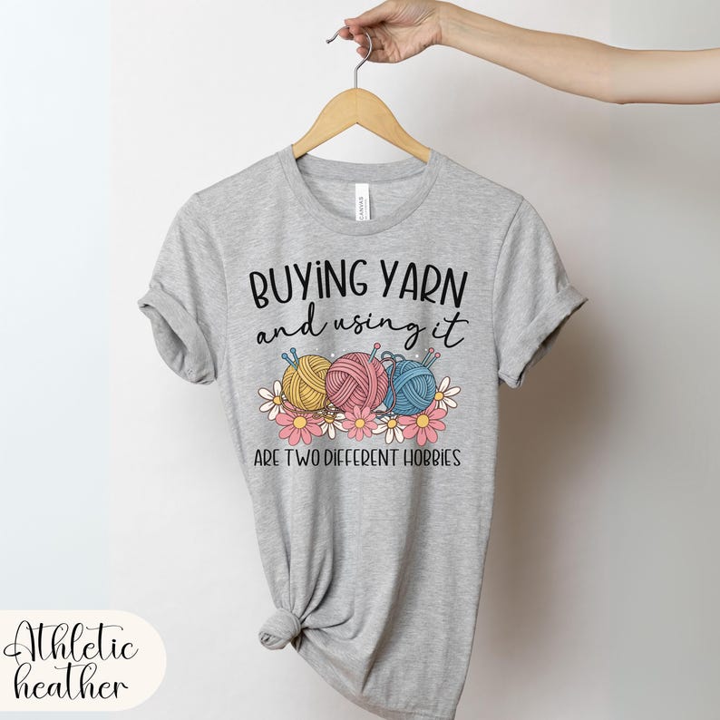 Puede incluir: Camiseta gris jaspeado con el texto "Buying Yarn and using it are two different hobbies". El gr&aacute;fico presenta tres ovillos de lana en amarillo, rosa y azul, con flores rosas y blancas. La camiseta est&aacute; colgada de una percha de madera.