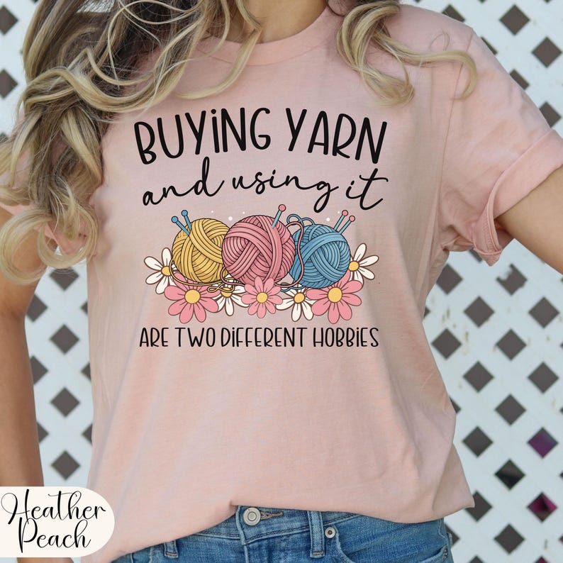 Puede incluir: Camiseta rosa claro con el texto "BUYING YARN and using it ARE TWO DIFFERENT HOBBIES". El dise&ntilde;o presenta tres ovillos de lana en amarillo, rosa y azul, con margaritas.