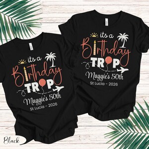 May include: Two black t-shirts with the text "its a Birthday TROP" in coral and white. The design includes a sun, palm tree, balloon, and airplane. The text "Maggie's 50th St Lucia - 2026" is also present.