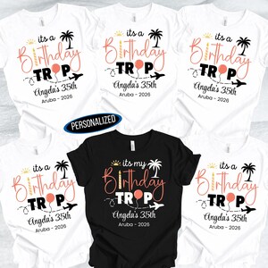 May include: White and black t-shirts with a birthday travel theme. The shirts feature the text "its a Birthday Trip" with a palm tree, airplane, and balloon graphic. The text "Angela's 35th Aruba - 2026" is also included.