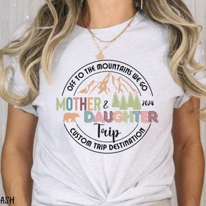 May include: White t-shirt with a colorful circle graphic that says "Off to the Mountains We Go Mother & Daughter Trip Custom Trip Destination 2024". The graphic features a mountain range, trees, and a bear.