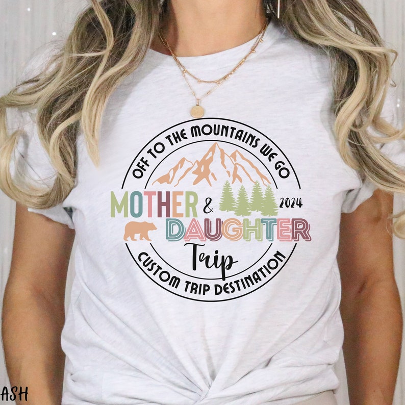 May include: White t-shirt with a colorful circle graphic that says "Off to the Mountains We Go Mother & Daughter Trip Custom Trip Destination 2024". The graphic features a mountain range, trees, and a bear.