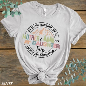May include: A light gray t-shirt with a circular design featuring a mountain range, a bear, and trees. The text "Off to the Mountains We Go" is above the image, and the text "Mother & Daughter Trip" is below the image. The text "Custom Trip Destination" is below the image. The year "2024" is on the right side of the design.