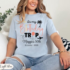 May include: Light blue t-shirt with the text "its my Birthday Trip" and "Maggie's 50th St Lucia - 2026". The design includes a candle, palm tree, balloon, and airplane. The shirt is light blue.