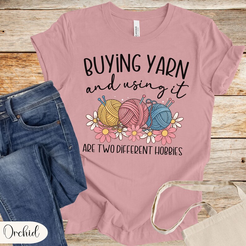 Puede incluir: Camiseta rosa con el texto "BUYING YARN and using it ARE TWO DIFFERENT HOBBIES". El dise&ntilde;o presenta ovillos de lana amarillos, rosas y azules, con flores blancas y rosas. Tambi&eacute;n se ven unos vaqueros azules y una bolsa de mano.