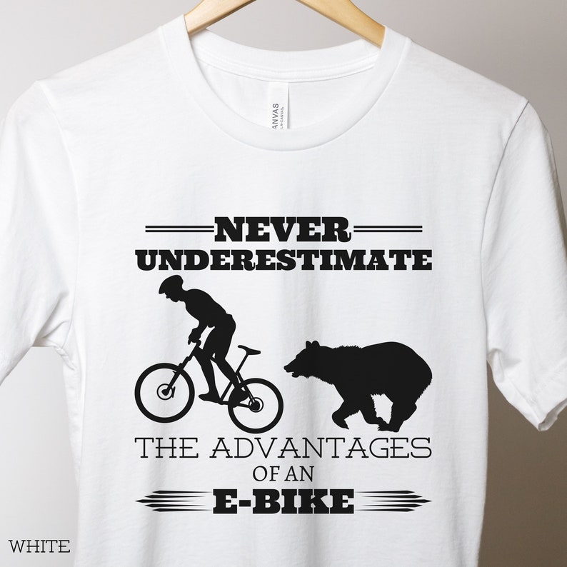 Op de afbeelding: Wit T-shirt met een zwart silhouet van een persoon op een fiets en een beer. De tekst luidt "Onderschat nooit de voordelen van een e-bike."