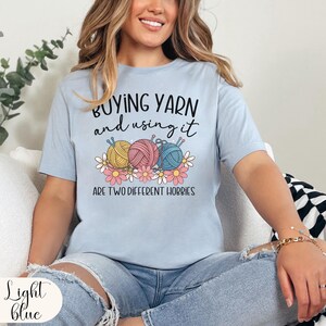 Puede incluir: Camiseta azul claro con el texto "BUYING YARN and using it ARE TWO DIFFERENT HOBBIES". La camiseta presenta un gr&aacute;fico de tres ovillos de lana en amarillo, rosa y azul, con margaritas.