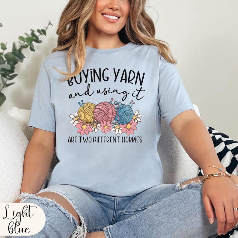 Puede incluir: Camiseta azul claro con el texto "BUYING YARN and using it ARE TWO DIFFERENT HOBBIES". La camiseta presenta un gr&aacute;fico de tres ovillos de lana en amarillo, rosa y azul, con margaritas.