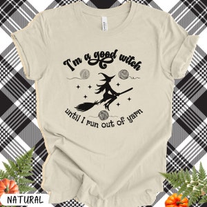 May include: A cream-colored t-shirt with the text "I'm a good witch until I run out of yarn" in black script. A witch on a broom is in the center, with yarn balls and stars. The shirt is a natural color.