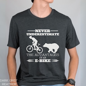 Op de afbeelding: Donkergrijs heather t-shirt met een wit grafisch ontwerp. De afbeelding toont een silhouet van een persoon die op een fiets rijdt en een beer. De tekst luidt "Onderschat nooit de voordelen van een e-bike."