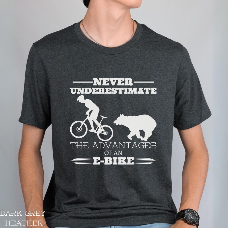 Op de afbeelding: Donkergrijs heather t-shirt met een wit grafisch ontwerp. De afbeelding toont een silhouet van een persoon die op een fiets rijdt en een beer. De tekst luidt "Onderschat nooit de voordelen van een e-bike."