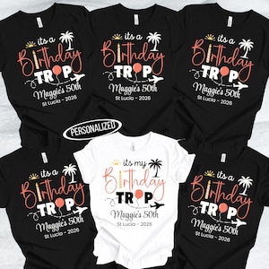 May include: A collection of black and white t-shirts with a birthday theme. The shirts feature the text "its a Birthday TROP" with a palm tree, airplane, and balloon graphic. The text "Maggie's 50th St Lucia - 2026" is also included.