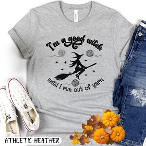 May include: Athletic heather gray t-shirt with the text "I'm a good witch until I run out of yarn." The design features a witch on a broom, yarn balls, and stars. The shirt is paired with blue jeans and white sneakers.