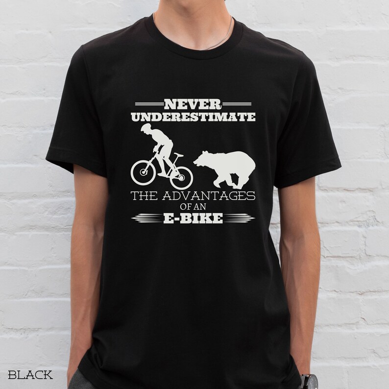 Op de afbeelding: Een zwart T-shirt met een wit grafisch ontwerp met de tekst "Onderschat nooit de voordelen van een e-bike". De grafiek bevat ook een silhouet van een persoon die op een fiets rijdt en een beer.