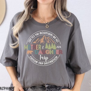 May include: A gray t-shirt with a white circle design featuring a mountain range, trees, a bear, and the text "Off to the Mountains We Go Mother & Daughter Trip 2024 Custom Trip Destination".