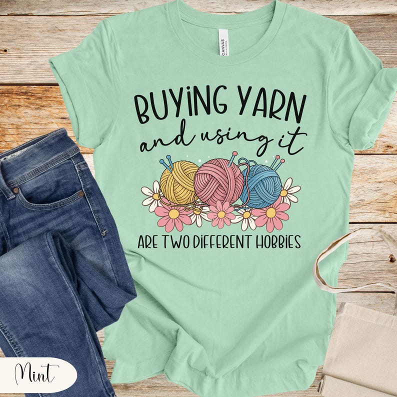 Puede incluir: Camiseta verde menta con el texto "COMPRAR HILO y usarlo SON DOS HOBBIES DIFERENTES." La camiseta presenta un gr&aacute;fico de tres ovillos de lana en amarillo, rosa y azul con agujas de tejer y flores.