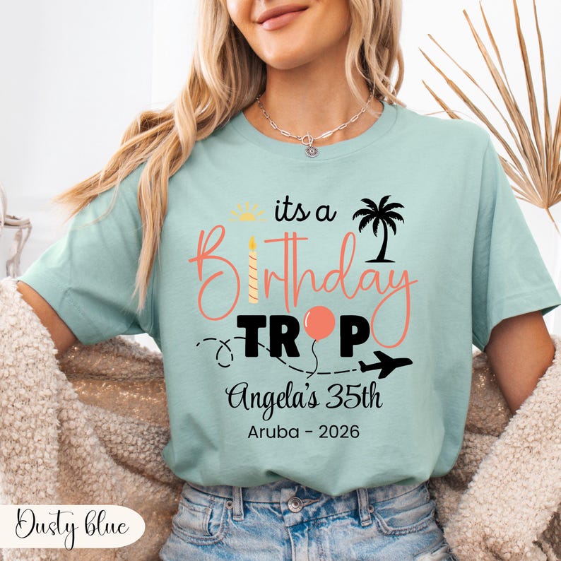 May include: Dusty blue t-shirt with the text "its a Birthday TR P" in coral and black. The design includes a palm tree, a candle, a balloon, and an airplane. The text "Angela's 35th Aruba - 2026" is also printed on the shirt.