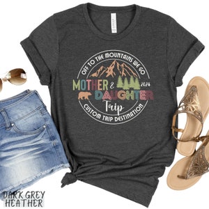 May include: Dark grey heather t-shirt with a circular graphic design featuring mountains, trees, a bear, and the text "Off to the mountains we go Mother & Daughter Trip Custom Trip Destination 2024".