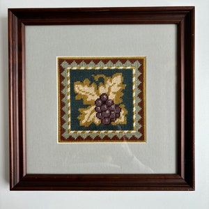 May include: A framed needlepoint picture depicting a bunch of purple grapes and leaves. The artwork is set against a dark teal background with a decorative border. The frame is dark brown wood, and the piece is mounted on a light grey mount.