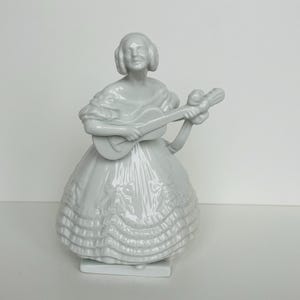 Puede incluir: Una figura de porcelana blanca de una mujer tocando la guitarra. La mujer lleva un vestido largo con volantes y tiene el pelo recogido en un moño. La figura está sobre una base rectangular.