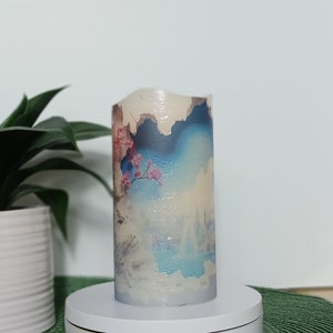May include: A cylindrical candle with a scenic design featuring mountains, clouds, and cherry blossoms. The candle has a white, textured top and is displayed on a white rotating stand. The color palette includes blue, pink, and white.