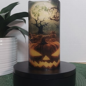 May include: A Halloween-themed candle with a jack-o'-lantern face and a spooky forest scene with a full moon. The candle is on a black circular base.