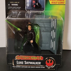 Op de afbeelding: Star Wars: The Power of the Force Luke Skywalker actiefiguur. De figuur zit in de originele verpakking, met een groen lichtzwaard en de tekst "Electronic Lightsaber Really Glows!". Inclusief het Rebel Alliance logo.