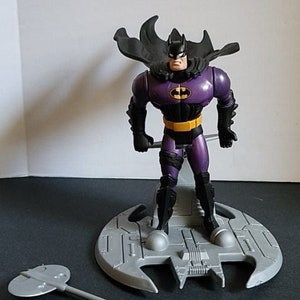 Pode incluir: Uma figura de ação do Batman de cor roxa e preta com uma capa e um batarang. A figura está em pé sobre uma base cinza em forma de morcego. A figura faz parte de um conjunto que inclui um batarang cinza.