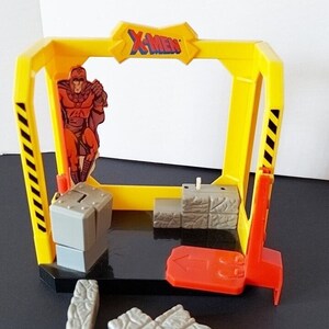 Op de afbeelding: Een gele plastic X-Men speelset met een rood en zwart Magneto-figuur. De speelset bevat een rode helling, grijze blokken en een geel frame met de tekst "X-Men" erop.