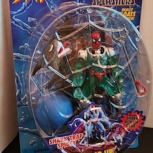 May include: Spider-Man action figure in packaging. The figure is in a clear plastic bubble, with a blue and red Spider-Man design. The packaging includes the text "Spider-Man", "Web Splashers", and "Shark Trap Spidey". Includes Shark Hunter Gear.