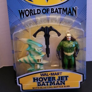 Pode incluir: Uma figura de ação do Batman verde com um cinto de ferramentas amarelo e uma capa preta, segurando um gancho de agarre preto. A figura é embalada com um brinquedo de jato de voo estacionário verde e branco. A embalagem apresenta o texto "World of Batman" e "Hover Jet Batman with Blasting Battle Sled".